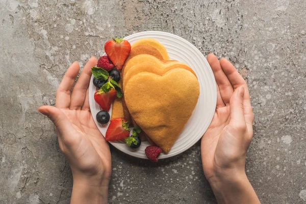 Vista parziale della donna che tiene il piatto con frittelle a forma di cuore e gustose bacche su una superficie di cemento grigio — Foto stock