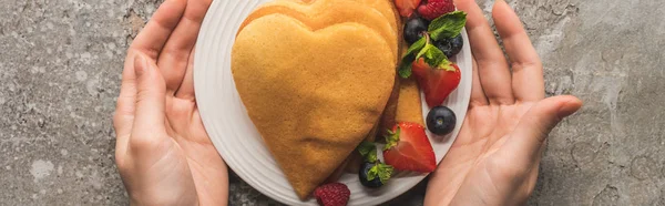 Partial view of woman holding plate with heart shaped pancakes and tasty berries on grey concrete surface, panoramic shot — Stock Photo