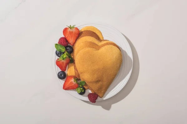 Vista superior de panqueques en forma de corazón con sabrosas bayas en el plato sobre fondo blanco — Stock Photo