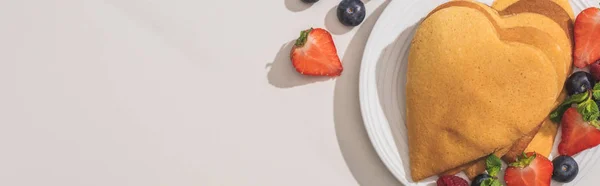Vista superior de panqueques en forma de corazón con sabrosas bayas en el plato sobre fondo blanco, plano panorámico - foto de stock
