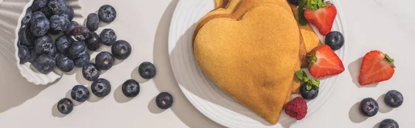 Draufsicht auf köstliche herzförmige Pfannkuchen in der Nähe verstreuter Blaubeeren auf weißem Hintergrund, Panoramaaufnahme — Stockfoto