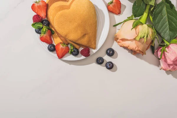 Vista superior de deliciosos panqueques en forma de corazón con bayas cerca de rosas sobre fondo blanco - foto de stock