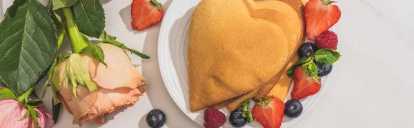 Vista superior de deliciosos panqueques en forma de corazón con bayas cerca de rosas sobre fondo blanco, plano panorámico - foto de stock