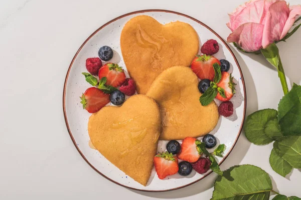 Vista superior de deliciosos panqueques en forma de corazón con bayas cerca de rosa sobre fondo blanco - foto de stock