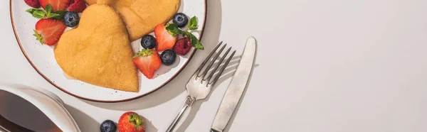 Top view of delicious heart shaped pancakes with berries near cutlery and maple syrup on white background, panoramic shot — Stock Photo