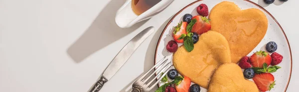 Vista superior de deliciosos panqueques en forma de corazón con bayas cerca de jarabe de arce y cubiertos sobre fondo blanco, plano panorámico - foto de stock