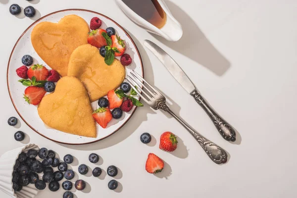 Vue de dessus de délicieuses crêpes en forme de coeur avec des baies près du sirop d'érable et des couverts sur fond blanc — Photo de stock