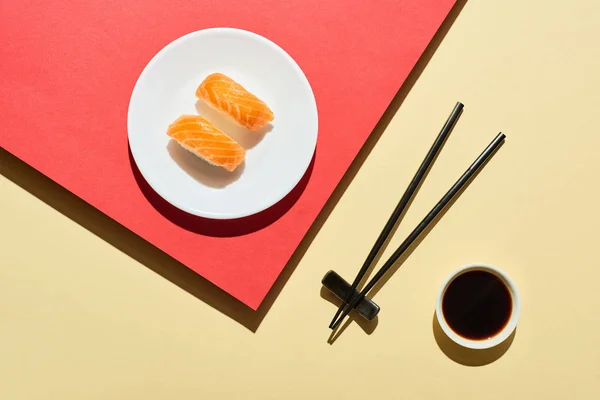 Vista superior de nigiri fresco con salmón cerca de salsa de soja y palillos en la superficie roja y beige - foto de stock