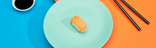 Fresh nigiri with salmon near soy sauce and chopsticks on blue and orange surface, panoramic shot — Stock Photo