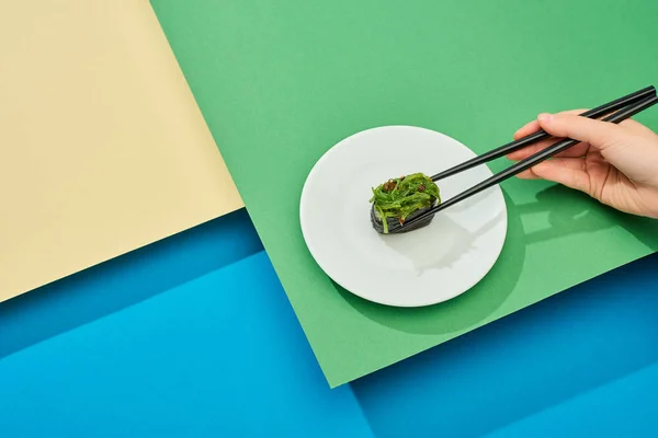 Vista recortada de la mujer comiendo nigiri fresco con algas marinas con palillos en la superficie multicolor - foto de stock