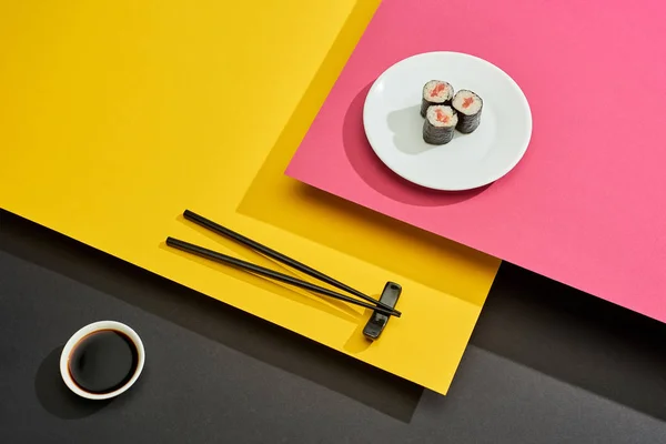 Fresh maki with salmon on plate near chopsticks and soy sauce on pink, yellow and black surface — Stock Photo