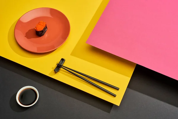 Fresh maki with red caviar on plate near chopsticks and soy sauce on pink, yellow and black surface — Stock Photo