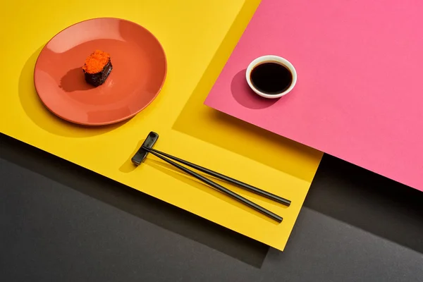 Fresh maki with red caviar on plate near chopsticks and soy sauce on pink, yellow and black surface — Stock Photo