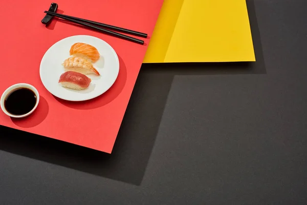 Nigiri frais au saumon, crevettes et thon près de sauce soja et baguettes sur surface rouge, jaune et noire — Photo de stock