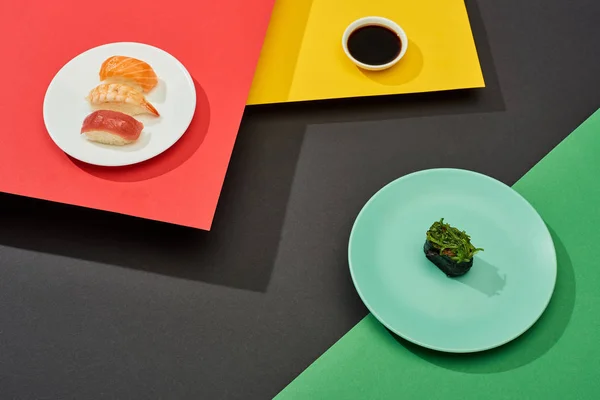 Fresh nigiri with salmon, shrimp and tuna near soy sauce and nigiri with seaweed on red, yellow, green and black surface — Stock Photo