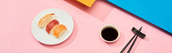 Fresh nigiri with salmon, tuna and shrimp near soy sauce and chopsticks on blue, pink, orange surface, panoramic shot — Stock Photo