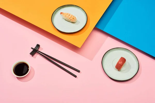 Свежий нигири с тунцом и креветками рядом с соевым соусом и палочками для еды на голубой, розовой, оранжевой поверхности — стоковое фото