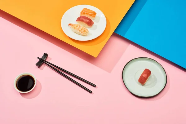 Fresh nigiri with salmon, tuna and shrimp near soy sauce and chopsticks on blue, pink, orange surface — Stock Photo