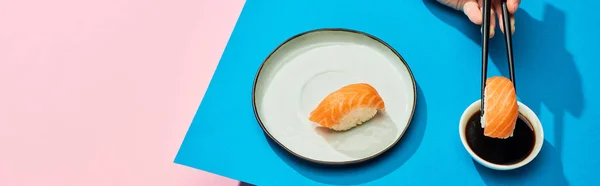 Cropped view of woman putting fresh nigiri with salmon into soy sauce on blue, pink background, panoramic shot — Stock Photo