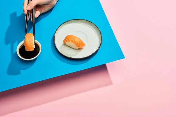 Cropped view of woman putting fresh nigiri with salmon into soy sauce on blue, pink background — Stock Photo