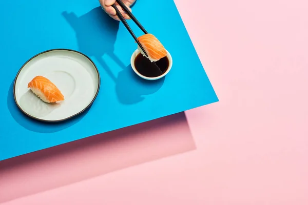 Cropped view of woman putting fresh nigiri with salmon into soy sauce on blue, pink background — Stock Photo