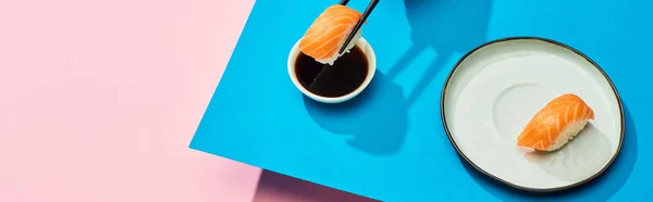 Fresh nigiri with salmon putting in soy sauce on blue, pink background, panoramic shot — Stock Photo