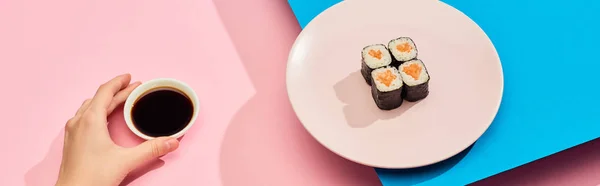 Cropped view of woman holding soy sauce near fresh maki with salmon on blue, pink background, panoramic shot — Stock Photo