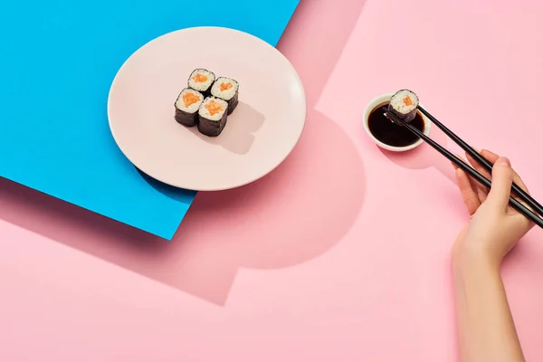 Vue recadrée d'une femme mangeant du maki frais avec du saumon près de la sauce soja sur fond bleu et rose — Photo de stock