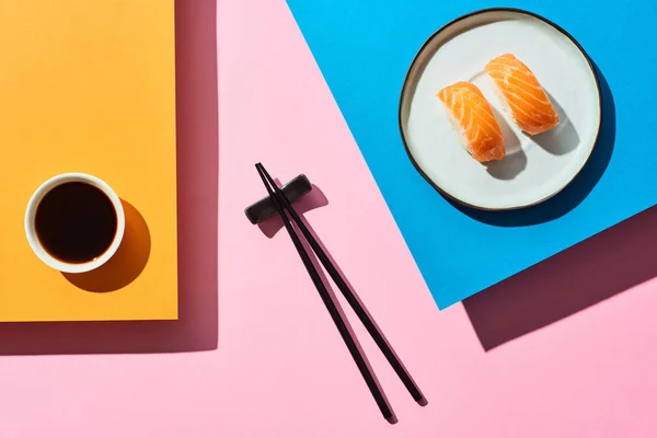 Top view of fresh nigiri with salmon near soy sauce and chopsticks on blue, pink, orange background — Stock Photo