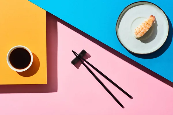 Top view of fresh nigiri with shrimp near soy sauce and chopsticks on blue, pink, orange background — Stock Photo