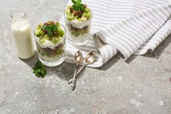 Muesli freschi con kiwi e yogurt su superficie di cemento grigio con tovagliolo rigato, bottiglia di latte e cucchiai — Foto stock