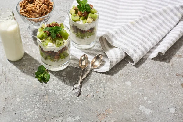 Fresh granola with kiwi and yogurt on grey concrete surface with striped napkin, bottle of milk, walnuts and spoons — Stock Photo