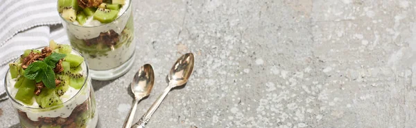 Fresh granola with kiwi and yogurt on grey concrete surface with striped napkin and spoons, panoramic shot — Stock Photo