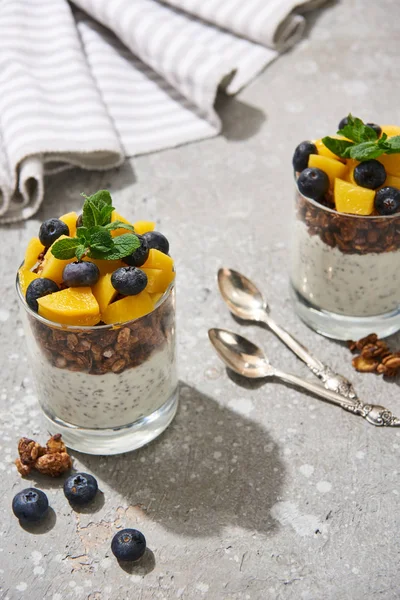 Selective focus of fresh granola with canned peach, blueberries and chia seeds on grey concrete surface with spoons, mint and napkin — Stock Photo