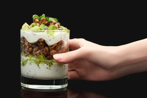 Vista recortada de mujer sosteniendo vidrio con granola fresca con kiwi y yogur aislado en negro - foto de stock