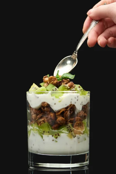Cropped view of woman holding spoon near fresh granola with kiwi and yogurt isolated on black — Stock Photo
