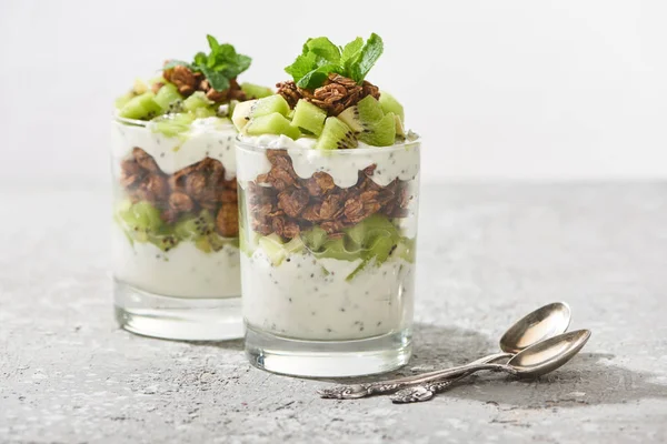 Messa a fuoco selettiva di muesli freschi con kiwi e yogurt vicino a cucchiai su superficie di cemento isolata su grigio — Foto stock