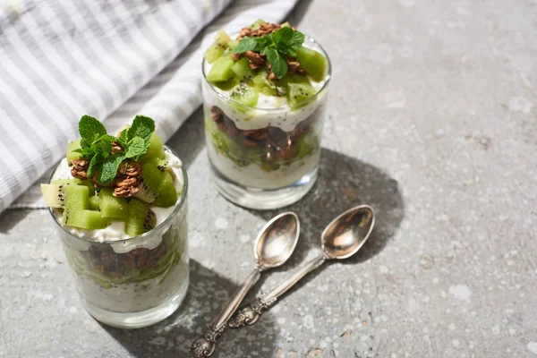 Frisches Müsli mit Kiwi und Joghurt auf grauer Betonoberfläche mit gestreifter Serviette und Löffeln — Stockfoto