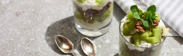 Frisches Müsli mit Kiwi und Joghurt auf grauer Betonoberfläche mit gestreifter Serviette, Löffeln, Panoramaaufnahme — Stockfoto