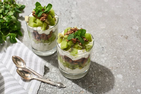 Fresh granola with kiwi and yogurt on grey concrete surface with striped napkin, spoons and mint — Stock Photo