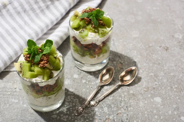 Fresh granola with kiwi and yogurt on grey concrete surface with striped napkin, spoons — Stock Photo