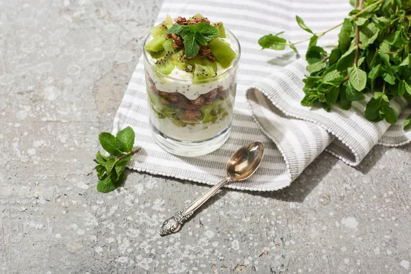 Fresh granola with kiwi and yogurt on grey concrete surface with striped napkin, spoon and mint — Stock Photo