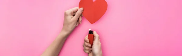 Vue recadrée d'une femme illuminant un cœur vide en papier rouge avec un briquet isolé sur une photo panoramique rose — Photo de stock