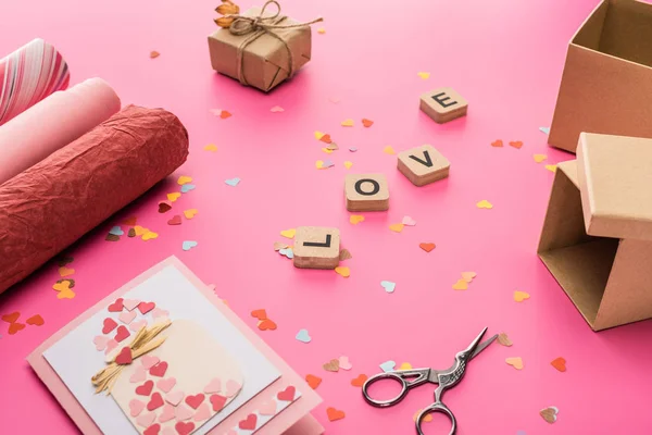Valentines confetti, scissors, wrapping paper, gift boxes, greeting card and love lettering on wooden cubes on pink background — Stock Photo