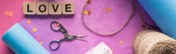 Vista superior da decoração dos namorados, tesoura, papel de embrulho, cordel, letras de amor em cubos de madeira sobre fundo rosa, tiro panorâmico — Fotografia de Stock