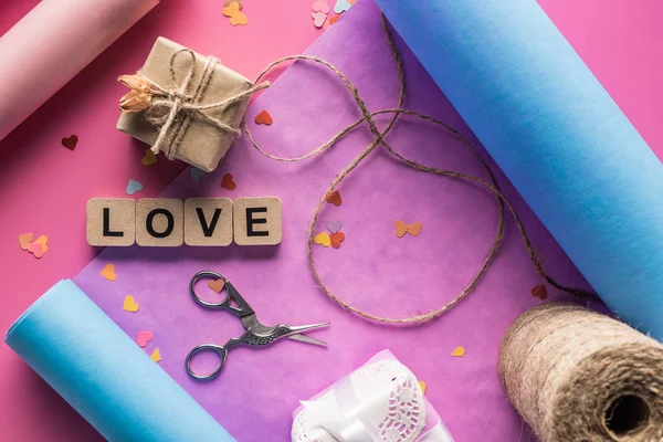 Vista superior de la decoración de San Valentín, tijeras, papel de envolver, cordel, cajas de regalo y letras de amor en cubos de madera sobre fondo rosa - foto de stock