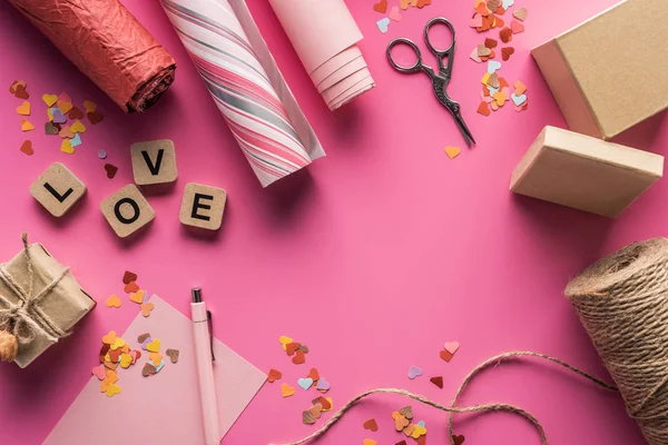 Ansicht von Valentinstag-Dekoration, Schere, Geschenkschachtel, Bindfaden, Geschenkpapier und Liebesschrift auf Holzwürfeln auf rosa Hintergrund — Stockfoto