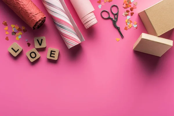 Ansicht von Valentinstag-Dekoration, Schere, Geschenkschachtel, Geschenkpapier und Liebesschrift auf Holzwürfeln auf rosa Hintergrund — Stockfoto