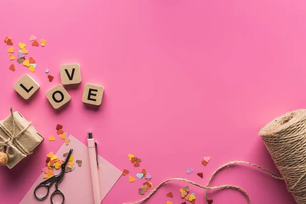 Vista superior de la decoración de San Valentín, tijeras, caja de regalo, hilo y letras de amor en cubos de madera sobre fondo rosa - foto de stock