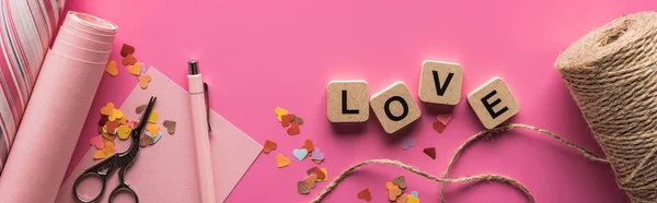 Top view of valentines decoration, scissors, wrapping paper, twine and love lettering on wooden cubes on pink background, panoramic shot — Stock Photo